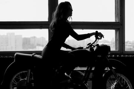 A silhouette of a woman with long hair sitting on a motorcycle. Black and white photoの写真素材