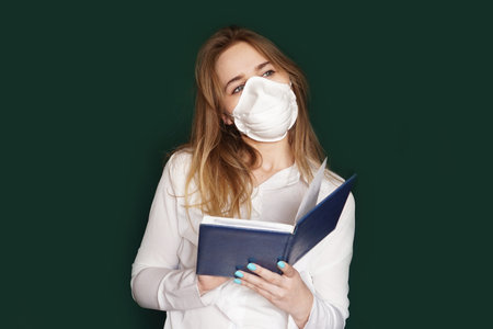 Young girl in a medical mask in a white office blouse with a notebook in hand on a green backgroundの写真素材