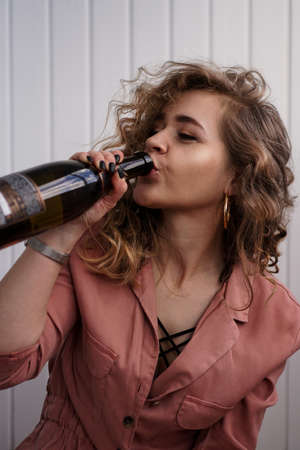 A young woman with curly hair drinks from a bottle of champagne. She is indoors on a white backgroundの写真素材