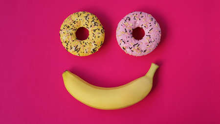 Two donuts and a banana lie on the pink surface, forming an abstraction of the emoticon - smile. Taken from the top pointの写真素材