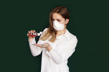 Young blonde girl in a white blouse and a mask uses an antiseptic on a green backgroundの写真素材