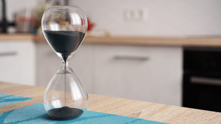 Closeup of hourglass on a white wooden table with defocus kitchen on background. Concept of time to...