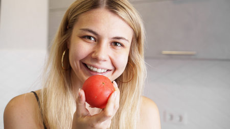 Young happy woman holding tomato and smiling on background of modern white kitchen. Healthy food...