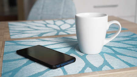 Clean white mug with handle and smart phone on blue table behind blurred kitchen backgroundの写真素材