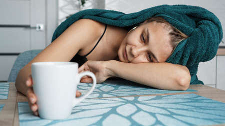 Tired woman sleeping on the table of the kitchen at breakfast. Beautiful girl in a towel on hair. Hand reaches for the mug. Lazy sleepy morningの写真素材