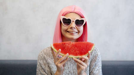 Indoor shot of young woman with piece of watermelon in hand, adorable female smiling at camera. Woman wears pink wig and heart-shaped glassesの写真素材