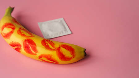Unpacked condom and red lipstick on a yellow banana on pink studio background. Contraceptives, prevention of venereal disease, preventing pregnancyの写真素材