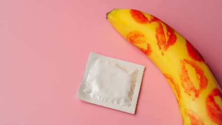 Unpacked condom and red lipstick on a yellow banana on pink studio background. Contraceptives, prevention of venereal disease, preventing pregnancyの写真素材
