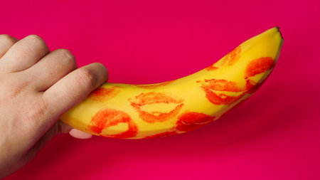 Woman hand holding banana with red lipstick markings on a pink background