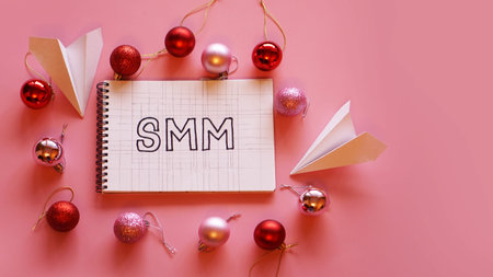 The inscription SMM in a notebook on a pink festive background. Red and pink Christmas balls, paper planes aroundの写真素材
