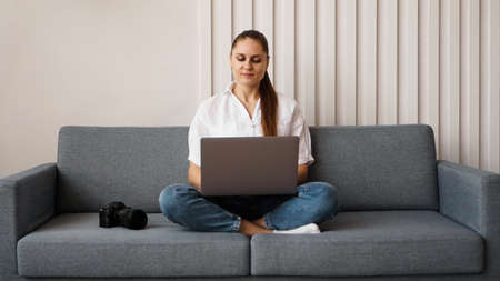 Happy young woman using laptop at home. On the couch next to the woman is a camera. Photographer retouches photographs at home.の写真素材