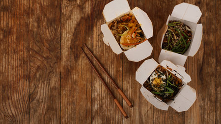 Noodles with pork and vegetables in take-out box on wooden table. Asian food delivery. Food in paper containers on wooden backgroundの写真素材