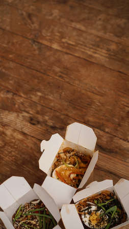 Noodles with pork and vegetables in take-out box on wooden table. Asian food delivery. Food in paper containers on wooden background. Vertical photoの写真素材