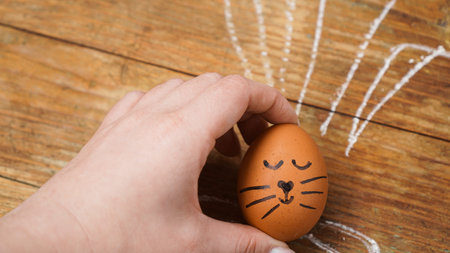 A female hand holds an egg with a cute face. The ears are drawn with chalk