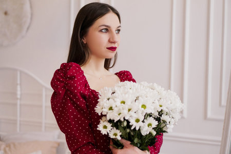 Beautiful brunette in a red dress with a bouquet of daisies at homeの写真素材