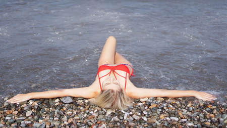 A beautiful blonde in a red swimsuit sunbathes on a pebble beach near the seaの写真素材