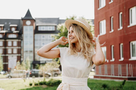 A beautiful blonde walks through a European city. Woman in white dress and straw hat, she smiles and happyの写真素材