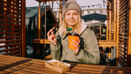 Woman eating tasty french fries and drinking tea or coffee in outdoor cafe. Woman at an urban...