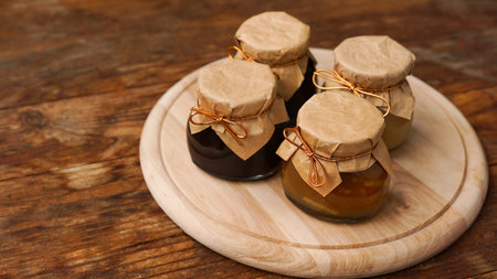 Four glass jars with different kinds of jam on a round cutting board on wooden table. Homemade jam,...
