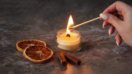 A woman hand holds a long match to light a homemade candle with the aroma of orange and cinnamon. Cozy atmosphere and aromatherapyの写真素材