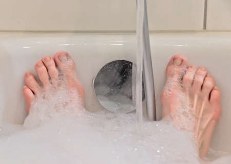 A bathtub in a bathroom filled with warm water and foam, only the feet of a person are shownの写真素材