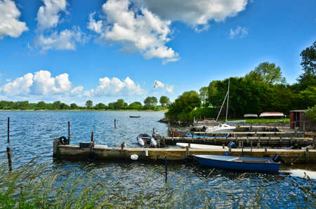 A beautiful coastline at a lake in Germany, Schlei, with a tiny boat at a pier in front of beautiful blue sky and white cloudsのeditorial素材