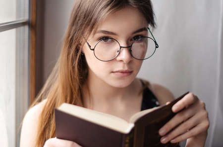 Beautiful girl sitting at the window, holding a book in his hands and looking at the cameraの写真素材