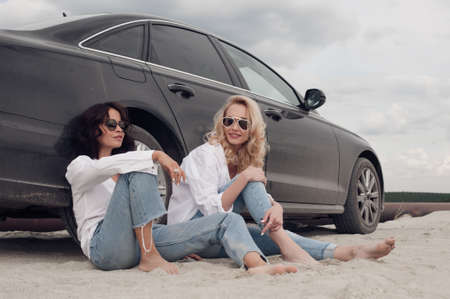 Two girls with glasses sitting on the beach near the car and talkの写真素材