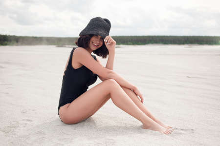 Beautiful brunette in a black bathing suit sitting on the beach and laughs looking at youの写真素材