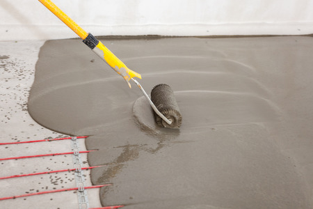 Electric floor heating system installation in new house. Worker align cement with rollerの写真素材