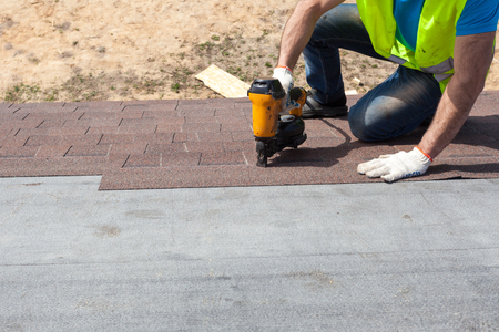 Roofer builder worker with nailgun installing Asphalt Shingles or Bitumen Tiles on a new house under constructionの写真素材