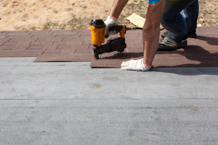 Roofer builder worker with nailgun installing Asphalt Shingles or Bitumen Tiles on a new house under constructionの写真素材