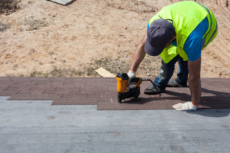 Roofer builder worker with nailgun installing Asphalt Shingles or Bitumen Tiles on a new house under constructionの写真素材