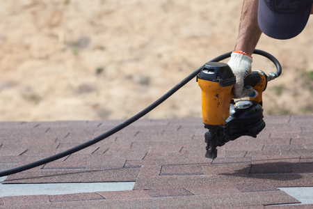 Roofer builder worker with nailgun installing Asphalt Shingles or Bitumen Tiles on a new house under construction