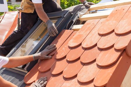 Hands of roofer laying tile on the roof. Installing natural red tile. Roof with mansard windows.の写真素材