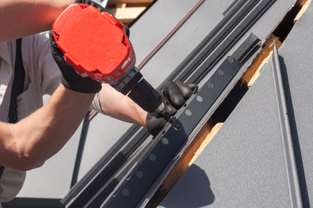 Worker on a roof with electric drill installing metal tile on wooden houseの写真素材