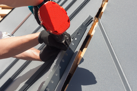 Worker on a roof with electric drill installing metal tile on wooden houseの写真素材