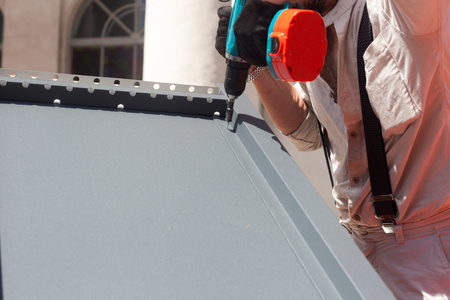 Worker on a roof with electric drill installing metal tile on wooden houseの写真素材