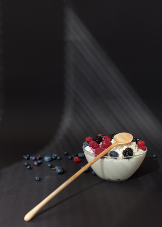 Fresh dairy product cottage cheese with berries in glass bowl with wooden spoon on black table.の写真素材