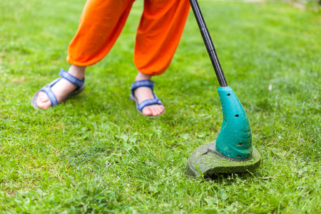 Gasoline lawn trimmer mows juicy green grass on a lawn on a sunny summer day. Close-up selective focus image. Garden equipment. Young woman mowing the grass with a trimmer.の写真素材