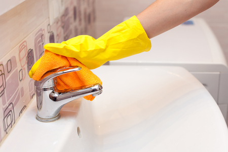 Female hands with yellow rubber protective gloves cleaning water tap with orange cloth.の写真素材