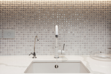 Modern designer chrome water tap over stainless steel kitchen sink. Interior of bright white kitchen. Water is poured through an open tapの写真素材