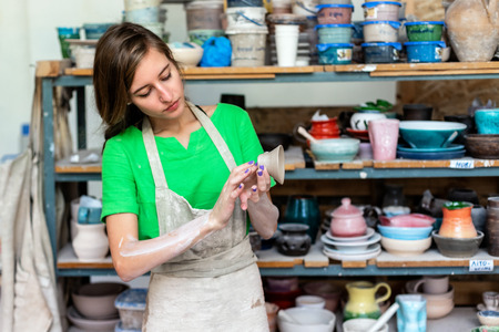 Master class concept. Gorgeous nice adorable good-looking lady in her casual green t-shirt workwear she stand indoor workspace against wooden shelf with utensil stuff background with clay cup in handの写真素材
