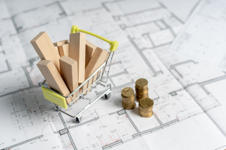 High and low price concept. High angle above top view small miniature object of shopping cart full of sawmill construction materials standing on table with stack of gold coins indoor light roomの写真素材
