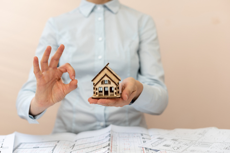Insure your home! Confident unrecognizable sale agent lady in her formal wear sit inside bright workstation. She show ok-sign on fingers hold small house in hand present new comfort flat apartmentの写真素材