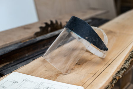 Photo of transparent protective face mask lying on the stack of lumber with blurred table saw on...