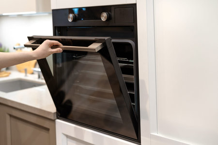 Woman hand opening door of new modern built-in oven in white kitchen cabinet