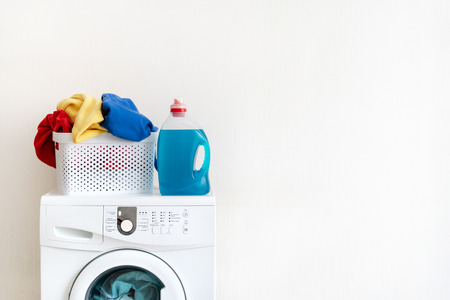 Preparation to laundry process. White washing machine with gel soap and basket of clothes on top standing isolated inside light flat interior with copy space for textの写真素材