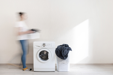 Time for laundry. Attractive lady going close to white washing machine with basket of clothes in hands. Laundromat standing inside bright apartment interiorの写真素材