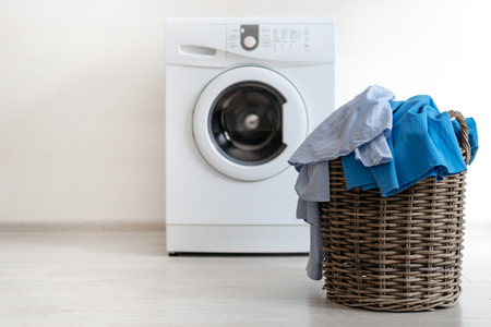 Laundry basket with pastel blue shirt and dress standing near white washing machine inside apartment with light interiorの写真素材